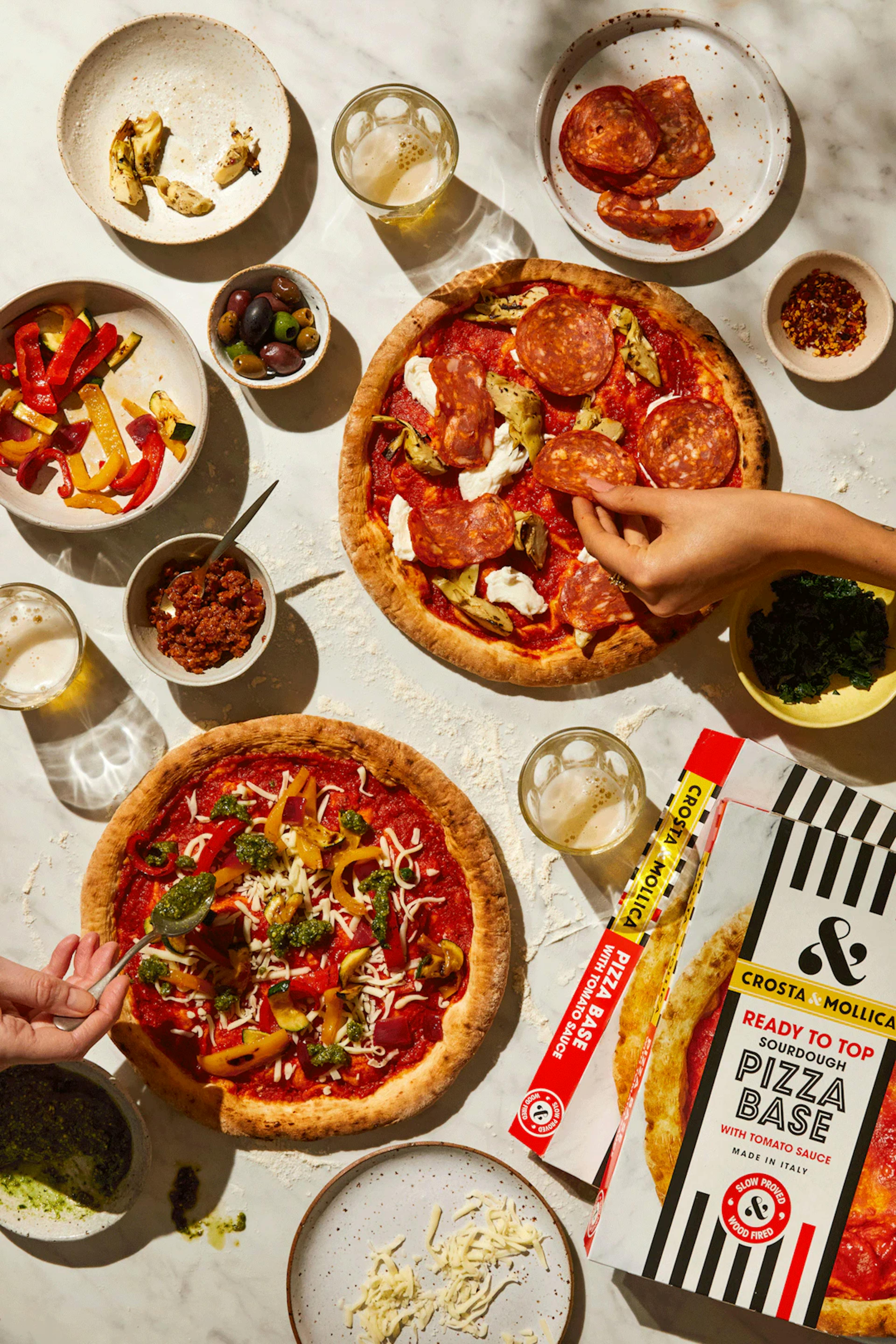 Various Crosta Mollica products laid out on a table. A hand adds a slice of Pepperoni to a Pizza.