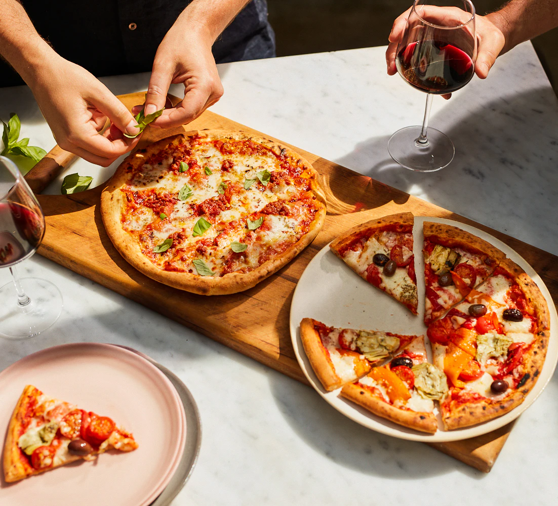 Two pizzas are on a board, two pairs of hands are reaching over them.
