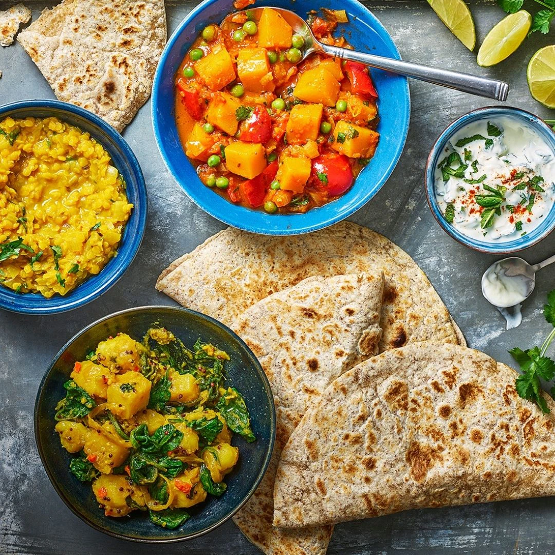 A bowl of saag aloo, one of parka dal and another of vegetable curry are shot from above. Organic Wholeblend Piadina are scattered between the bowls.