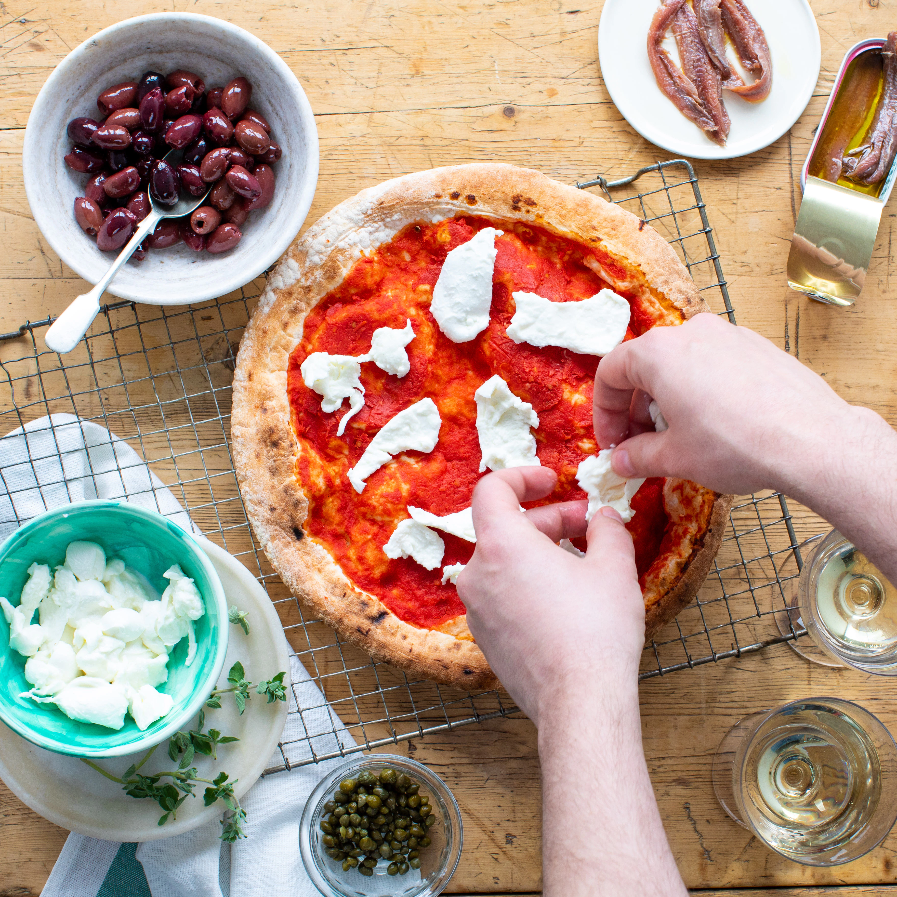 A pair of hands are placing pieces of shredded mozzarella on a pizza base which has tomato sauce on it. The pizza base is on a wire cooling rack with small bowls of ingredients surrounding it. 