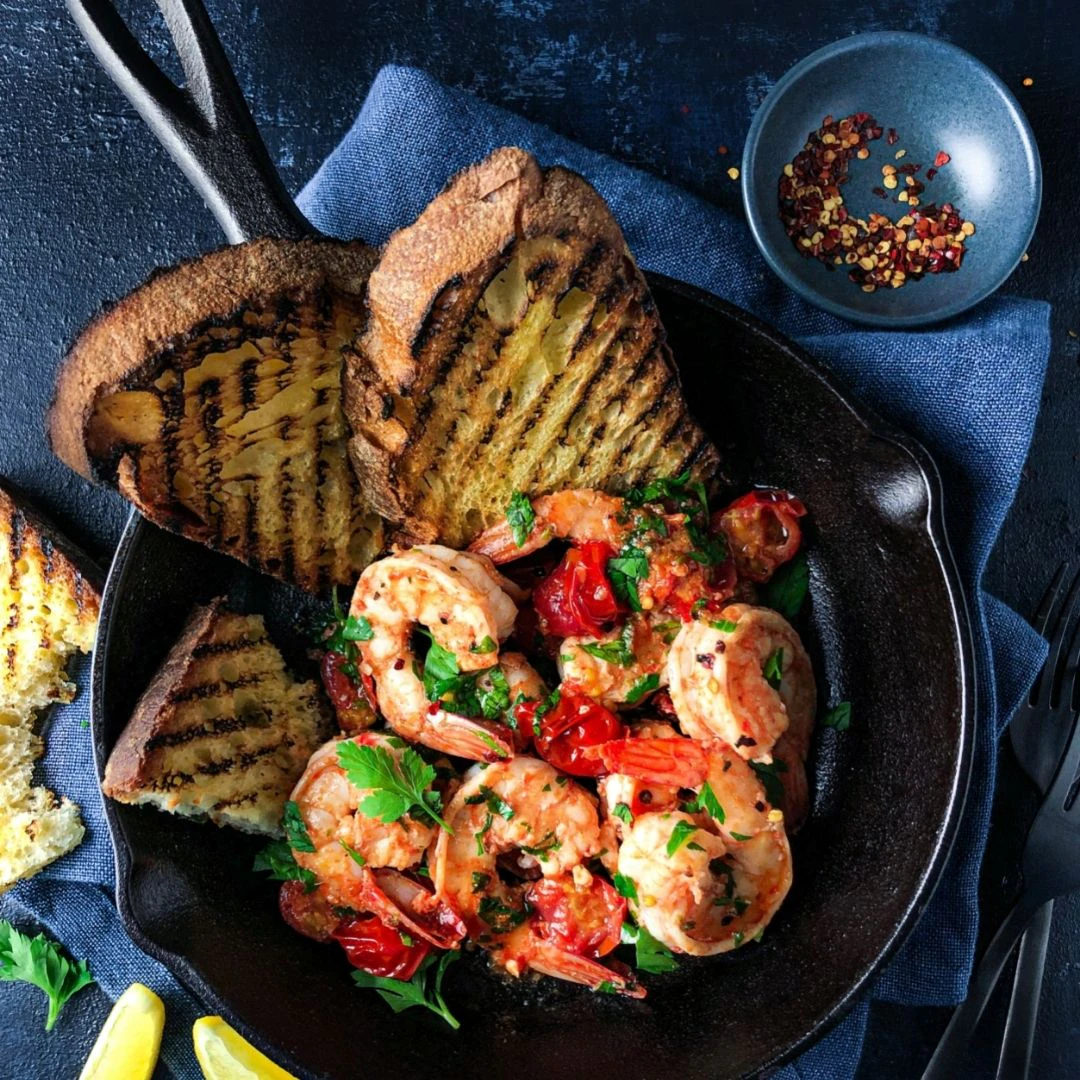A bowl with king prawns in garlic butter, three slices of charred bread are on the side of the bowl.