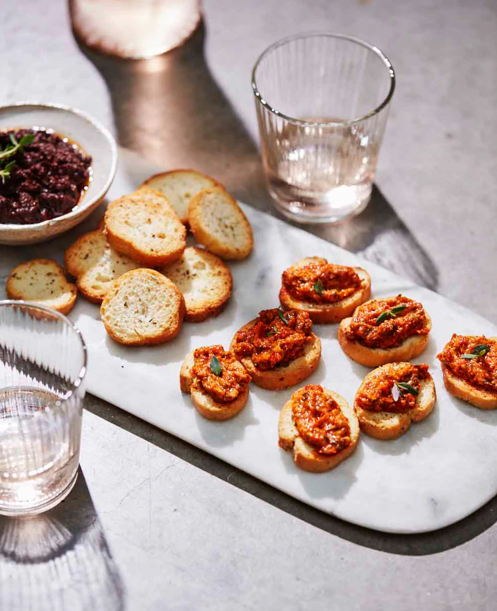 Oregano crostini topped with a red dip are on a tray next to three glasses.