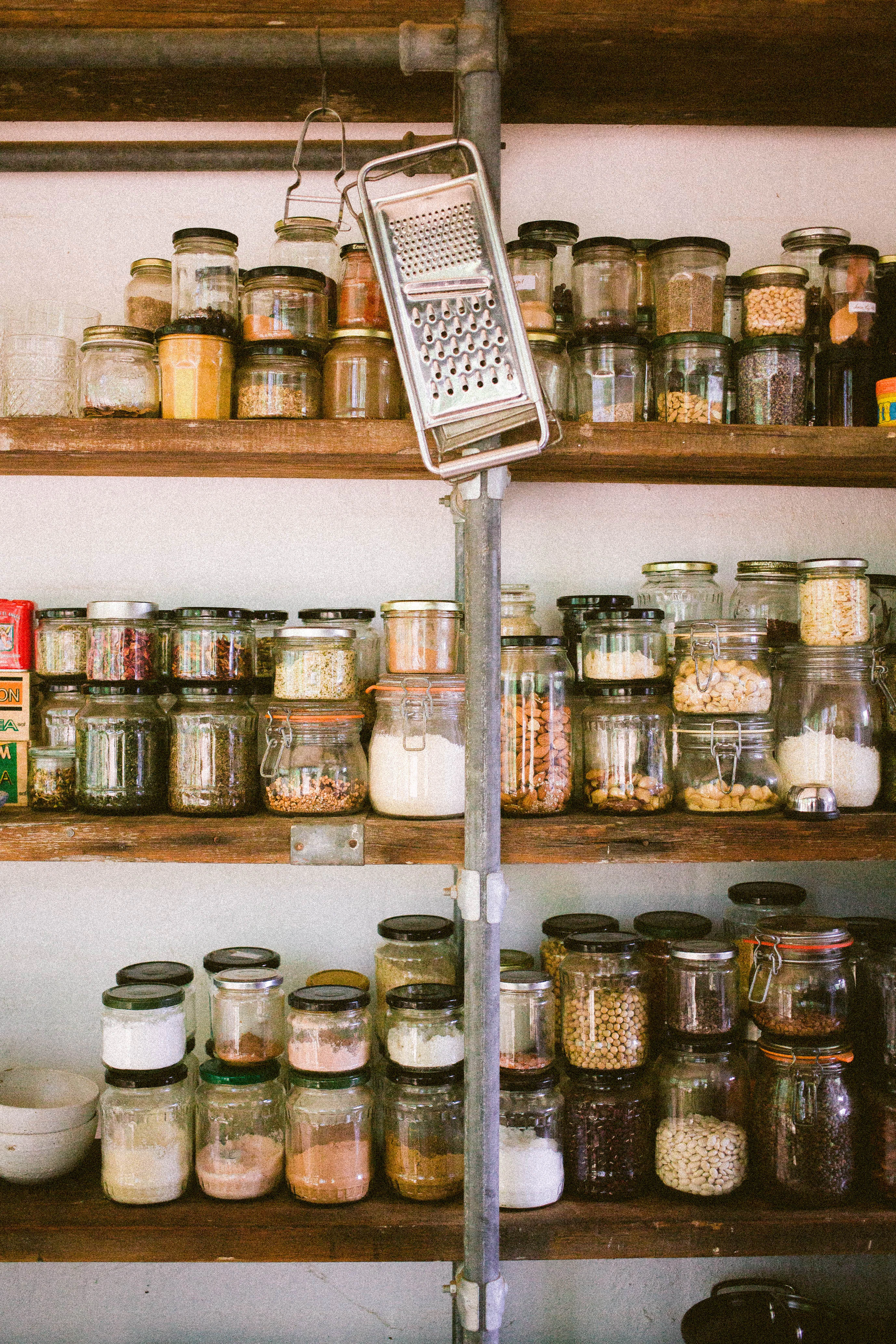 Three shelves with glass jars of ingredients on it, a grater hangs from a shelf.