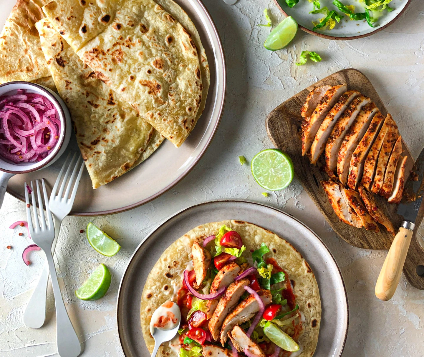 Three plates are in view, one has folded Piadina on it with a pot of pickled red onions, on a different plate is an assembled lime and chilli chicken piadina, the third plate has sliced chicken breast on it.