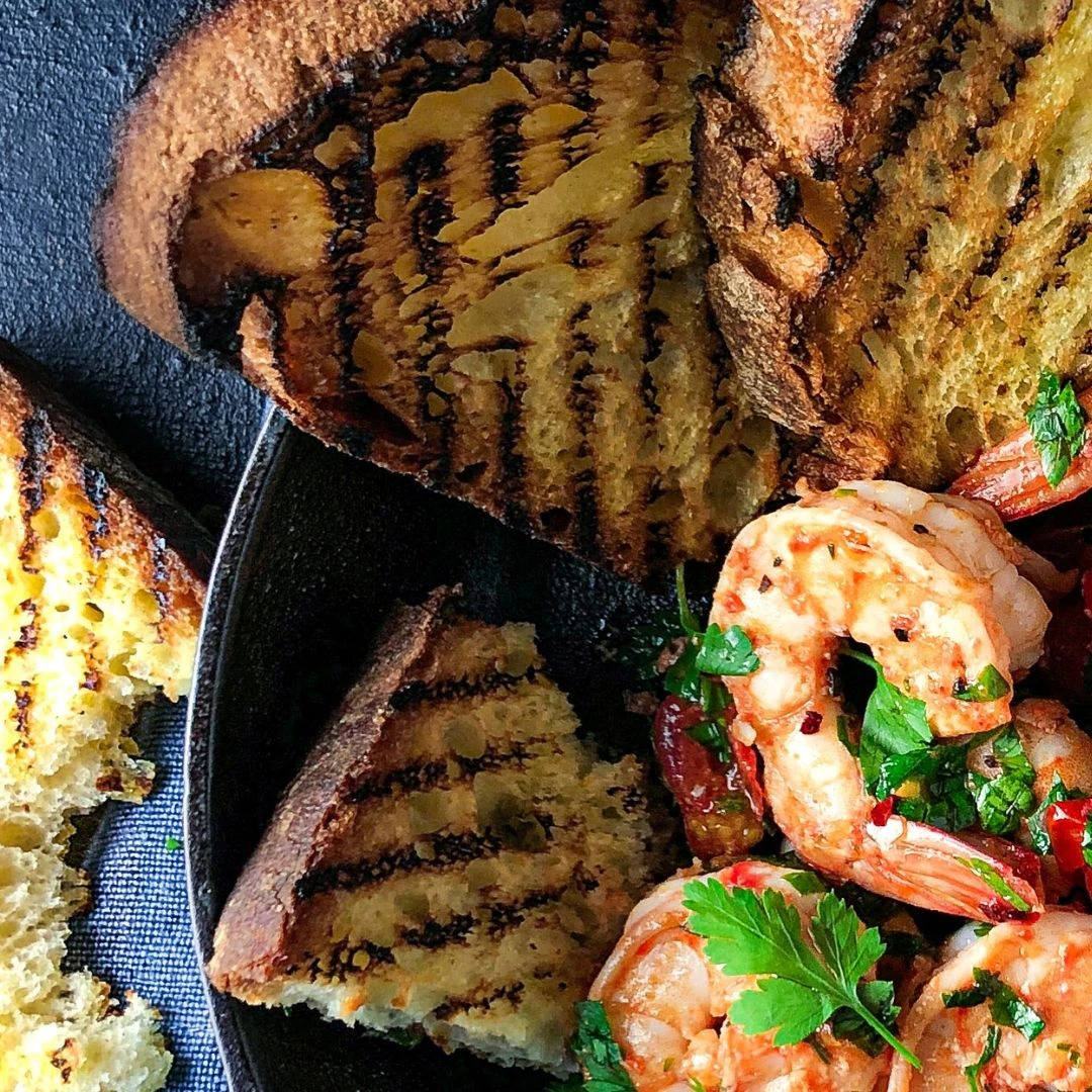 Three slices of charred Pane Pugliese are in a bowl next to some prawns.
