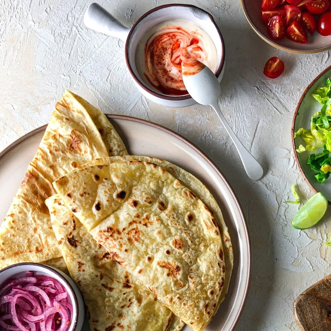 Three Piadina are folded on a large plate next to a pot of pickled red onions. Two bowls of salad and a ramekin of sirachia mayonnaise are next to them.