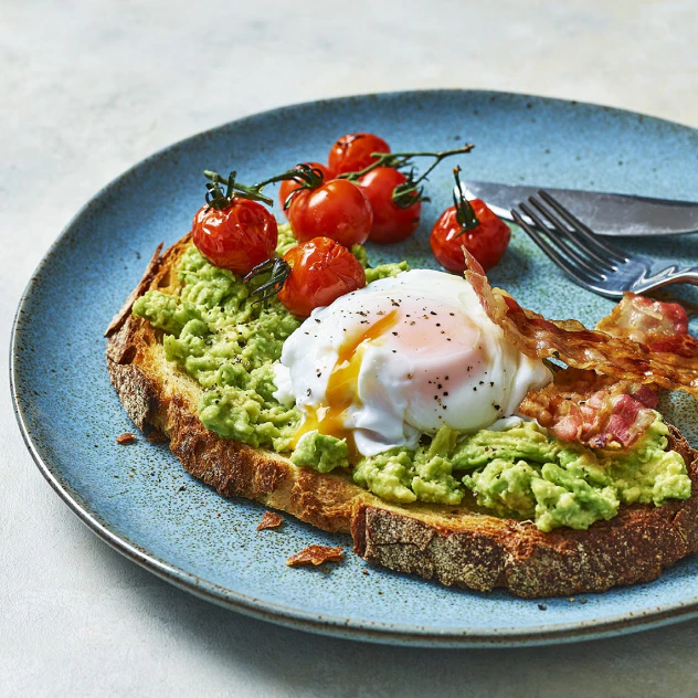 A plate has a slice on Pane Pugliese toast with avocado, a poached egg and pancetta on top. Roasted tomatoes are also on the plate, along with a knife and fork.