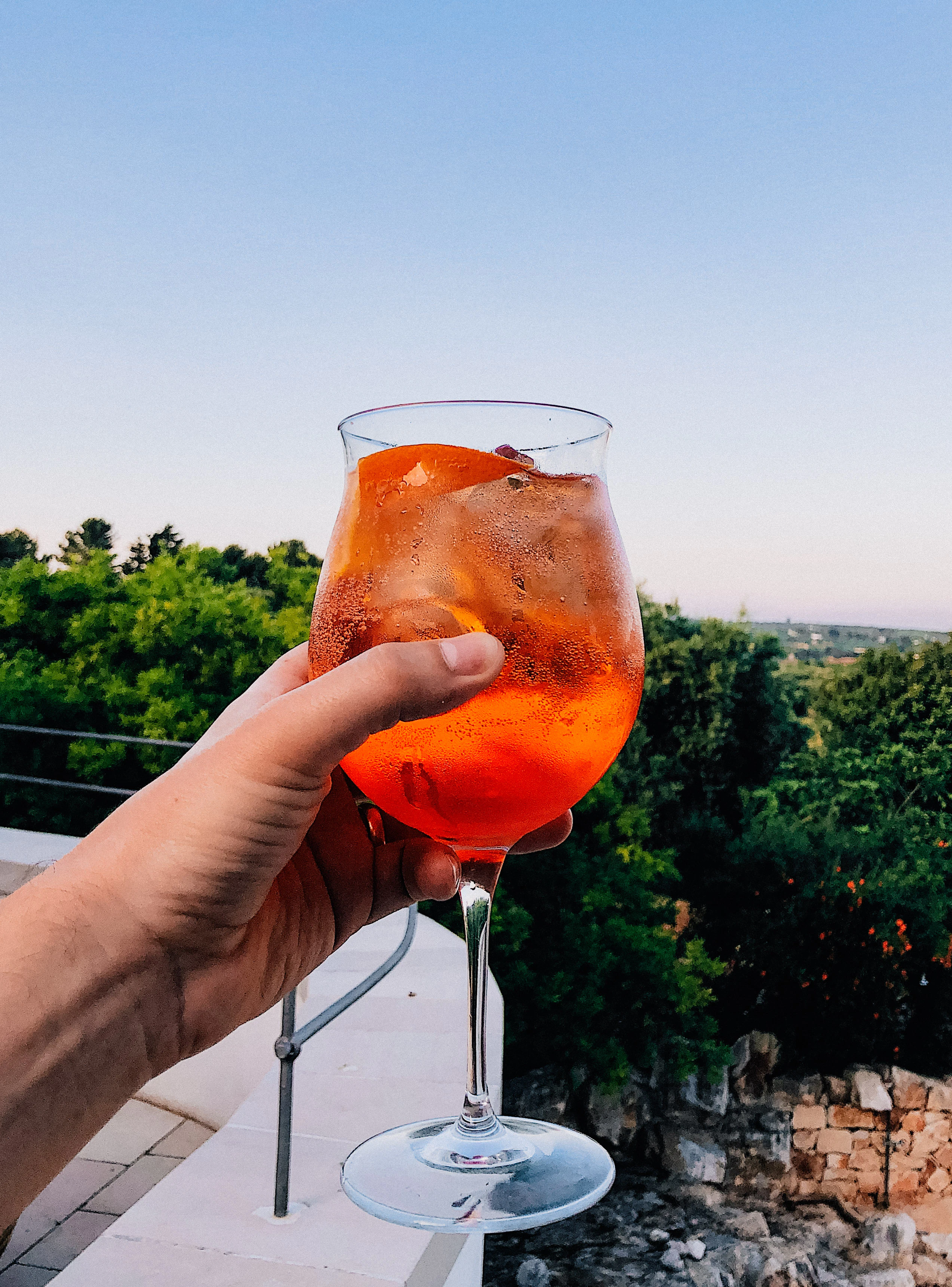 A hand holds out an Aperol spritz, in the background is an Italian landscape.