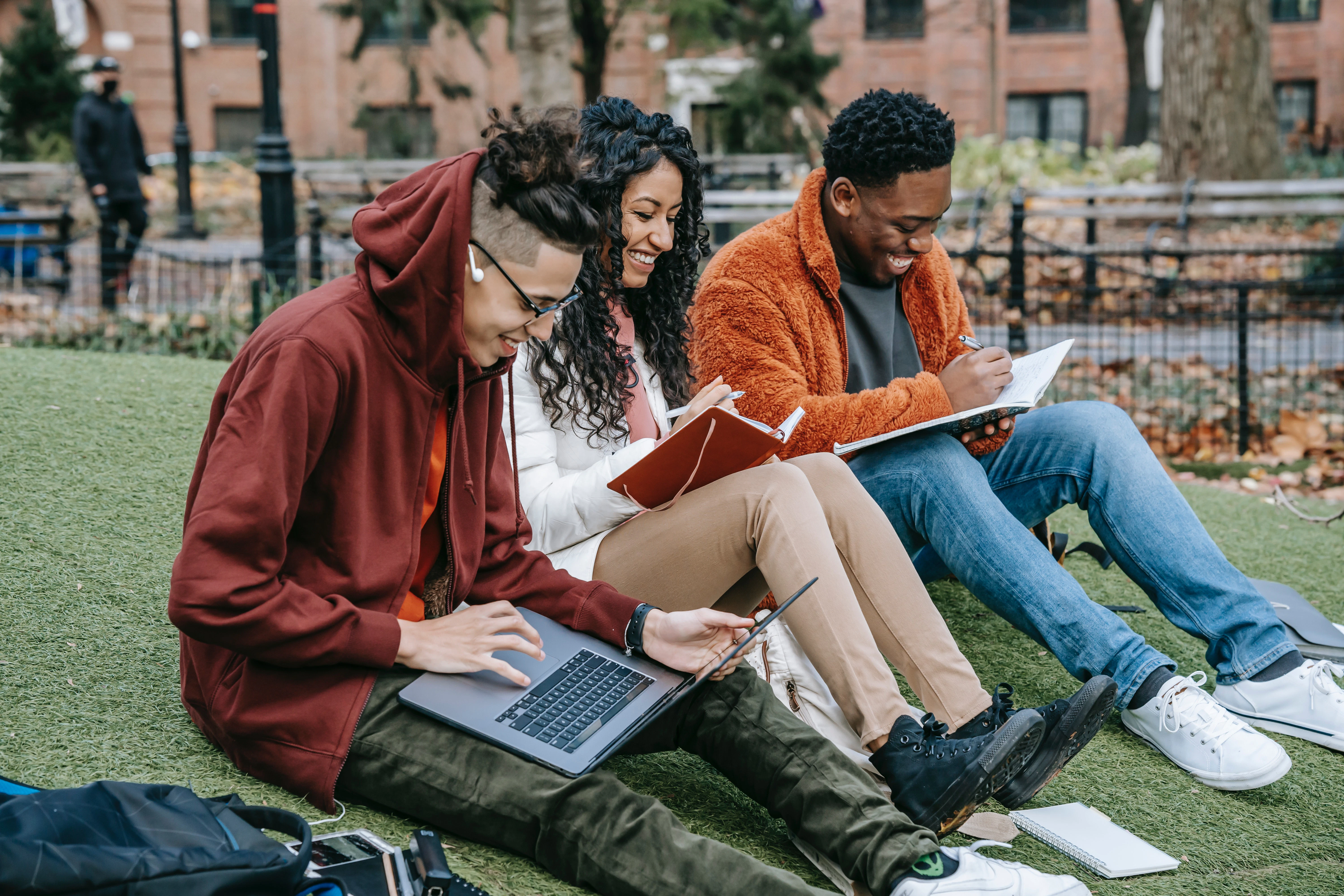 A picture of a three college students with their books and laptops open, sitting in the grass.