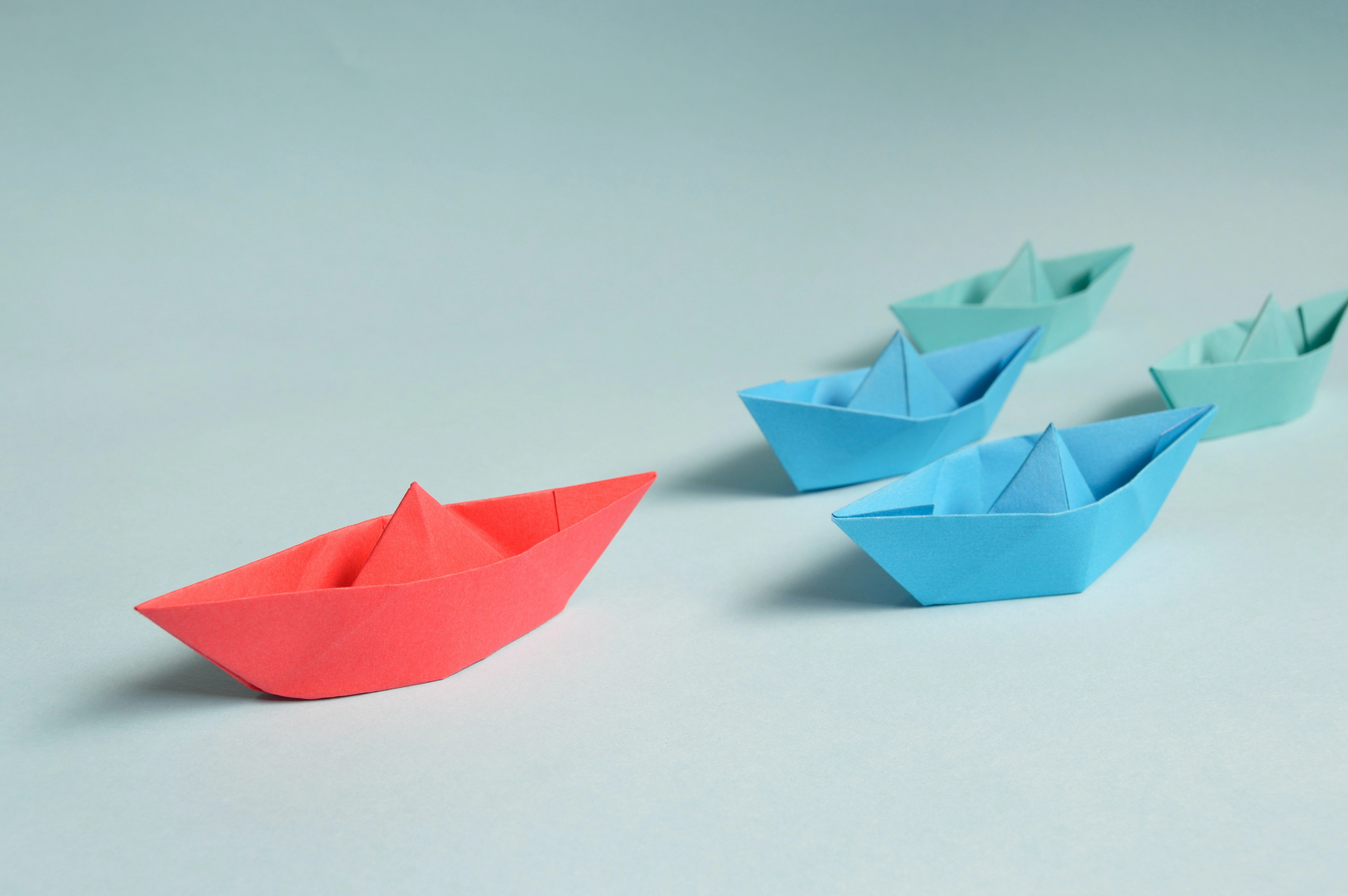 a photo of five paper boats on a white surface, with a red paper boat in lead.