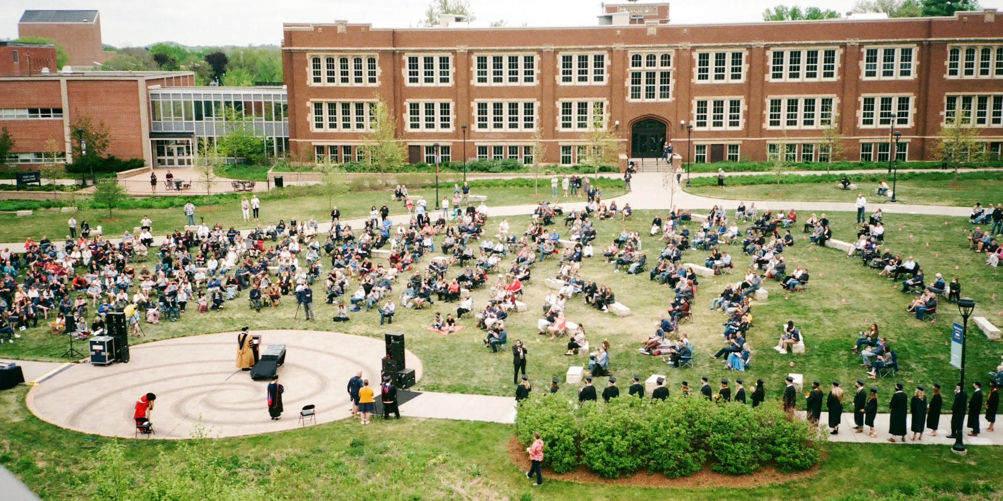 Students assembled outdoors on a college campus, listening to a speaker.